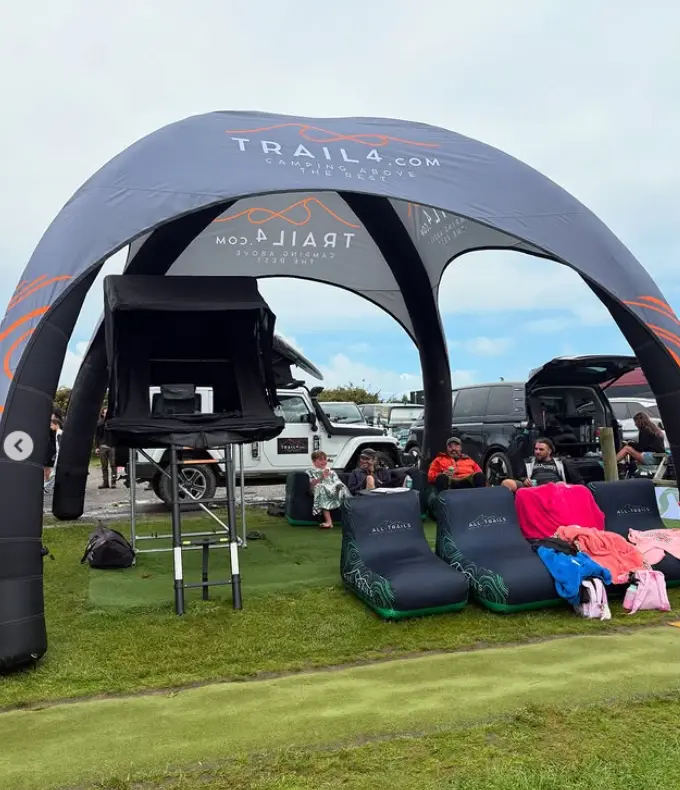 A wide shot of a branded "TRAIL4.COM - CAMPING ABOVE THE REST" inflatable dome tent set up at an outdoor event, featuring a rooftop tent on a stand and people relaxing on comfortable inflatable "ALL TRAILS" chairs on a grassy field.